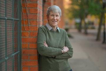 Stephanie 奈斯比特 in a green sweater, leans against a brick wall with her arms folded.